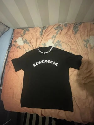 Svart t-shirt med tryck 'AESTHETIC' - Svart t-shirt med vit text 'AESTHETIC' på bröstet och tryck i nacken. Klassisk passform och rund hals. Perfekt för dig som gillar streetwear och vill ha en enkel men snygg look.