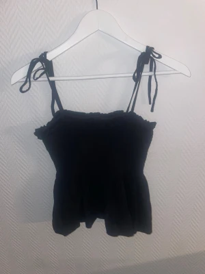 Svart linnetopp  - Snygg svart linnetopp med smockad överdel och tunna axelband som knyts på axlarna. Toppen har en lätt volangkant upptill och nedtill, vilket ger en söt och trendig look. Perfekt att matcha med jeans eller kjol.