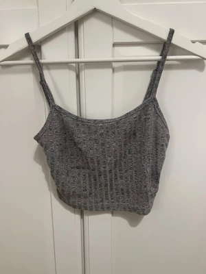 Grå ribbad linnetopp - Säljer en enkel och stilren grå ribbad linnetopp med smala axelband. Toppen har en croppad passform och är perfekt för varma dagar eller som lager under en skjorta. Materialet känns mjukt och stretchigt.