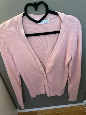 Ljusrosa ribbad kofta med knappar - Säljer en ljusrosa ribbad kofta med v-ringning och knäppning framtill. Koftan har långa ärmar och är tillverkad i ett mjukt, stretchigt material. Perfekt att slänga över en topp för en chill look.