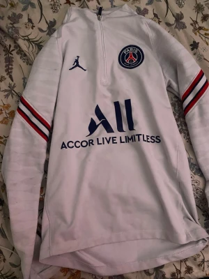 Vit PSG träningströja långärmad - Snygg vit långärmad träningströja från PSG med blå logga och text, röda och blå ränder på ärmarna samt halv dragkedja fram. Tillverkad i funktionsmaterial som passar perfekt till fotbollsträning eller sport. Paris-tryck på ena ärmen och Visit Rwanda på ryggen.