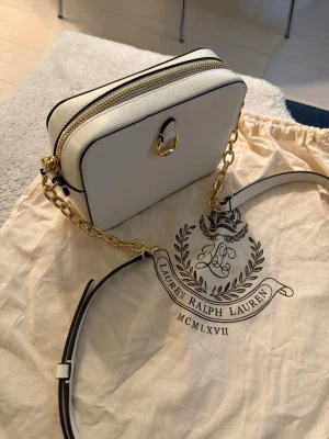 Vit handväska från Lauren Ralph Lauren - Säljer en stilren vit handväska från Lauren Ralph Lauren i fyrkantig modell. Väskan har guldfärgade metalldetaljer, kedja och justerbar axelrem i läder. Dragkedja upptill och elegant präglad struktur. Perfekt accessoar för en trendig look.