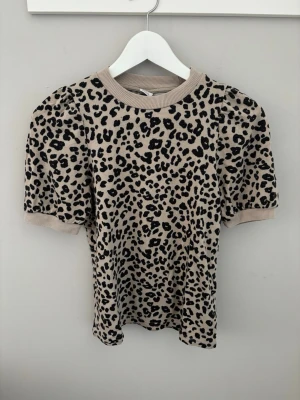 Leopardmönstrad topp  - Säljer en beige topp med svart leopardmönster och puffiga korta ärmar. Ribbad kant vid halsringning och ärmslut ger en snygg kontrast. Perfekt för dig som vill sticka ut lite extra! Materialet känns mjukt och stretchigt.