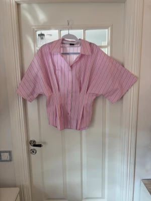 Rosa randig skjorta med kort ärm - Säljer en oversized rosa skjorta med vita och svarta ränder. Skjortan har korta ärmar, klassisk krage och knappar framtill. Perfekt för dig som gillar en avslappnad och trendig stil. Materialet känns som bomull.