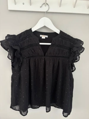 Svart volangblus  - Snygg svart blus från Gina med volangdetaljer på axlarna och diskret prickigt mönster i tyget. Blusen har en luftig passform, v-ringning och är tillverkad i ett tunt, transparent material som ger en lätt och flowy känsla.