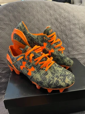 Under Armour camo fotbollsskor  - Snygga fotbollsskor från Under Armour med camouflagemönster i grönt och svart samt starka orange detaljer. Skorna har orange snörning, dobbar och logga. Tillverkade i syntetmaterial för bra grepp och komfort på planen. Perfekta för dig som vill sticka ut på matchen!