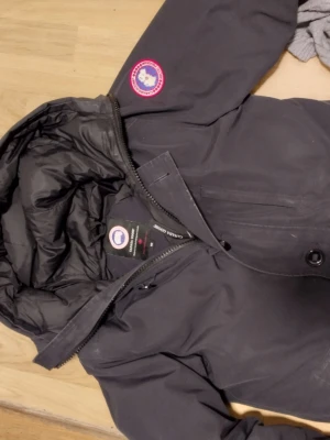 Svart dunjacka från Canada Goose medium - Säljer en svart beg dunjacka från Canada Goose med huva och klassisk logotyp på ärmen. Jackan har dragkedja och knappar framtill samt flera praktiska fickor. Perfekt för kalla vinterdagar tack vare det varma dunfodret... Beg skick. Fel fri men beg. Normal passform. Medium.