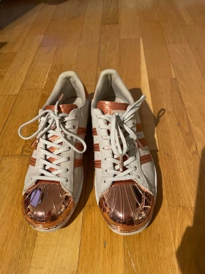 Adidas Superstar med kopparfärgade detaljer - Säljer ett par Adidas Superstar sneakers i vitt med metallic kopparfärgade tåhättor och detaljer. Skorna har klassiska tre ränder i koppar, vita snören och ovandel i mockaliknande material. Perfekta för dig som vill sticka ut med en unik twist på en ikonisk modell.
