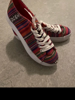 Färgglada mönstrade sneakers tygskor randiga - Säljer ett par unika sneakers med färgglatt randigt och etno-inspirerat mönster i rött, lila, grönt och svart. Skorna har vita snören och en tjock vit sula. Perfekta för dig som vill sticka ut med din stil!