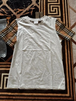 Vit Burberry t-shirt med rutiga ärmar - Snygg vit t-shirt från Burberry med klassiska beige, svarta och röda rutiga kortärmar. T-shirten har en rak passform och rund halsringning. Perfekt för dig som vill ha en stilren look med en touch av lyx.