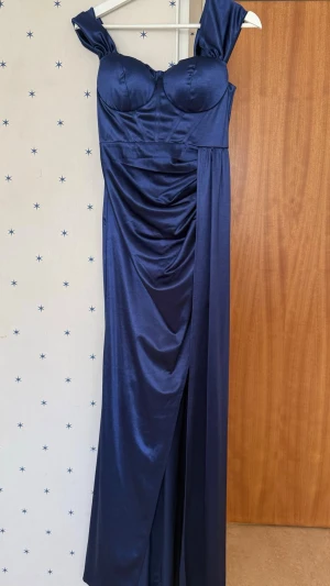 Marinblå långklänning med drapering - Marinblå jumpsuit i glansigt material med breda axelband och markerad byst. Draperad midja och vida ben ger en elegant och modern look. Perfekt för dig som vill sticka ut med stil. 