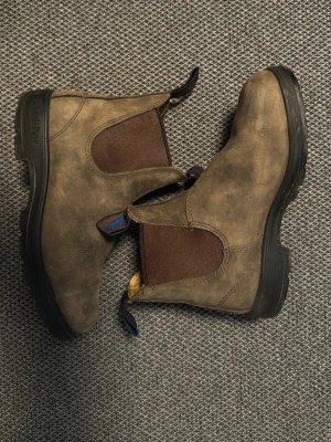 Bruna  boots från Blundstone - Säljer ett par klassiska Chelsea boots från Blundstone i brunt läder med elastiska sidopaneler och robust svart sula. Skorna har blå detaljer på dragflikarna och en tidlös, rundad tå. Perfekta för dig som gillar en avslappnad men stilren look.
