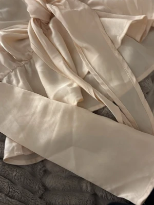 Krämvit blus med knytband - Snygg krämvit blus från Boohoo med långa ärmar och v-ringning. Blusen har ett dekorativt knytband framtill och är gjord i ett lätt, glansigt material som ger en elegant känsla. Perfekt att styla med jeans eller kjol för en trendig look.