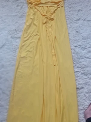Gul axelbandslös maxiklänning - Säljer en solgul axelbandslös långklänning från glance.. Klänningen har en bred smock upptill, knytband i midjan som du kan knyta på olika sätt. och ett mjukt, flowy tyg som ger en avslappnad känsla. Perfekt för varma dagar och sommarens alla tillfällen. Klänningen är aldrig använd så väldigt gott skick 