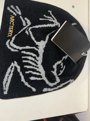 Svart mössa från Arc'teryx - Säljer en svart mössa från Arc'teryx med grått grafiskt mönster och gul logotyp på sidan. Mössan är stickad och har en tight passform som sitter snyggt på huvudet. Perfekt för dig som vill ha en stilren och sportig look.