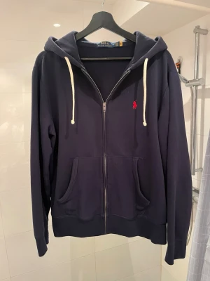 Mörkblå Polo Ralph Lauren zip - Mörkblå hoodie med dragkedja från Polo Ralph Lauren. Klassisk design med vit dragsko i huvan, två stora fickor framtill och röd broderad logga på bröstet. Tillverkad i mjukt bomullsmaterial, perfekt för chill dagar.