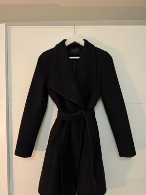 Svart kappa från VILA med skärp - Stilren figursydd svart kappa från VILA med knytskärp i midjan och bred krage. Kappan har dubbelknäppning framtill och två fickor på sidorna. Perfekt för dig som vill ha en klassisk och tidlös look under kalla dagar❤️