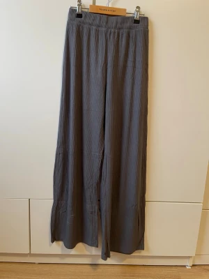 Grå vida mjukisbyxor med resårmidja - Säljer ett par grå mjukisbyxor med breda ben och ribbad struktur. Byxorna har resår i midjan och är tillverkade i ett mjukt, flowy material som ger en avslappnad vibe. Modellen är tänkt att vara kortare och sluta ovanför ankeln. Perfekta för chill dagar eller när du vill ha det extra bekvämt.