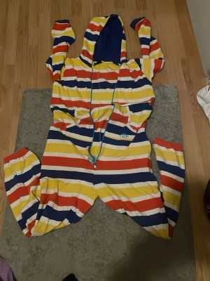 Randig onesie med huva - Färgglad onesie med breda ränder i gult, rött, vitt och mörkblått. Har huva med blått foder och dragkedja framtill. Perfekt för att chilla hemma i, supermjuk och skön passform. Långa ärmar och ben, samt fickor på sidorna.