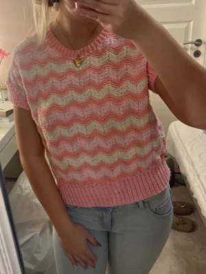 Rosa stickad topp med vågmönster - Säljer en söt stickad topp i rosa, vitt och ljusorange med vågigt mönster. Kortärmad modell med ribbstickad kant vid ärmar, hals och nederkant. Perfekt för dig som gillar pastell och retrovibbar. Materialet känns mjukt och luftigt.