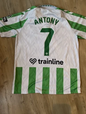Grönvit Real Betis matchtröja Hummel - Säljer en grönvit randig Real Betis matchtröja från Hummel med nummer 7 och namnet Antony på ryggen. Tröjan har klubbmärke på bröstet, sponsorloggor och klassisk krage. Perfekt för dig som älskar fotboll och vill sticka ut på planen. Säljer billigt pga defekt på höger arm