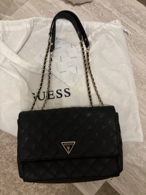 Svart handväska från Guess - Snygg svart handväska från Guess i quiltat konstläder med guldfärgade kedjedetaljer och topphandtag. Väskan har två mindre fack inuti samt ett större med dragkedja. Axelremmen kan bäras med båda handtagen och vara lite kortare samt med endast ett och bli längre. Finns några missfärgningar av smink på innertyget men inget som förstör annars är den i mycket bra skick för att vara väl använd