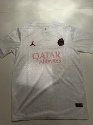 Vit PSG x Jordan matchtröja - Säljer en vit PSG x Jordan matchtröja med rosa Qatar Airways-tryck på bröstet och PSG-logga. Tröjan är tillverkad i Dri-FIT-material som andas och har ett subtilt mönster i tyget. Perfekt för fotboll eller träning.
