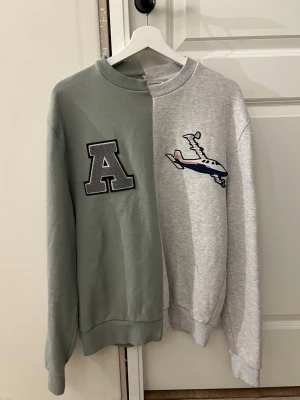 Axel Arigato tvåfärgad sweatshirt - Unik sweatshirt från Axel Arigato med tvådelad design i grönt och ljusgrått. Ena sidan har ett stort A-tryck och den andra ett broderat flygplan. Crewneck med ribbade muddar och avslappnad passform. Använd fåtal gånger 