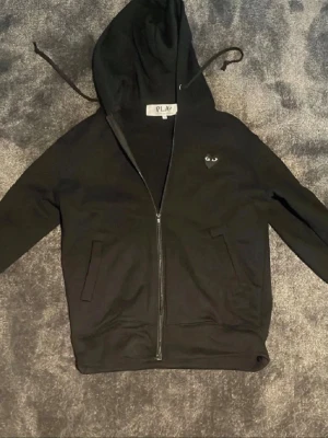 Svart hoodie från Comme des Garçons Play - Svart hoodie med dragkedja från Comme des Garçons Play. Har ett litet hjärta med ögon på bröstet och ett stort hjärta med ögon på ryggen. Utrustad med huva och snören samt två fickor framtill. Tillverkad i mjukt bomullsmaterial för en skön känsla.