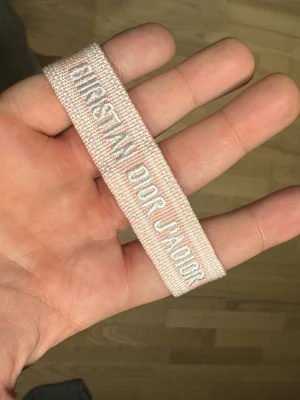 Christian Dior rosa tygarmband - Säljer ett snyggt rosa tygarmband från Christian Dior med texten 'Christian Dior J'Adior' i vitt. Armbandet har en vävd struktur och justerbara tofsar i ändarna för enkel passform. Perfekt accessoar för att lyfta din outfit med en exklusiv touch.