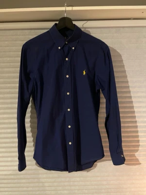 Marinblå skjorta från Ralph Lauren - Snygg marinblå skjorta från Ralph Lauren i slim fit med klassisk krage och knappar framtill. Den är i bra skick och säljs då den blivit liten. Pris 399.
