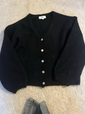 Svart kofta från Gina Tricot - Mysig svart kofta från Gina Tricot i ullblandning. Koftan har v-ringning, långa ärmar och knappar framtill. Perfekt att slänga över en topp för en enkel och snygg look.