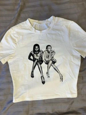 Vit croppad t-shirt med tryck Naomi Campbell & Kate Moss - Vit croppad t-shirt med ett svartvitt grafiskt tryck men en ikonisk bild på Naomi Campbell & Kate Moss. T-shirten har rund hals och korta ärmar. 💕