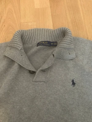 Grå stickad tröja från Polo Ralph Lauren - Säljer en stilren grå stickad tröja från Polo Ralph Lauren med ribbad krage och muddar. Tröjan har knappar vid halsen och den klassiska mörkblå loggan broderad på bröstet. Perfekt för dig som gillar tidlös och enkel design.