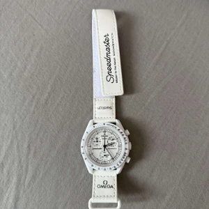 Vit Omega x Swatch Speedmaster moonwatch klocka - Säljer en vit Omega x Swatch Speedmaster klocka med kronograf och detaljer i svart. Klockan har vit boett och vitt armband i syntetmaterial med tydliga loggor på både urtavla och band. Stilig och modern design med klassiska Speedmaster-drag.