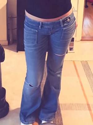 Blå bootcut jeans med slitningar - Säljer ett par ljusblå bootcut jeans med slitna detaljer vid knäna och framsidan. Jeansen har låg midja, vida ben och klassiska fickor fram och bak. Perfekta för en avslappnad och trendig look.
