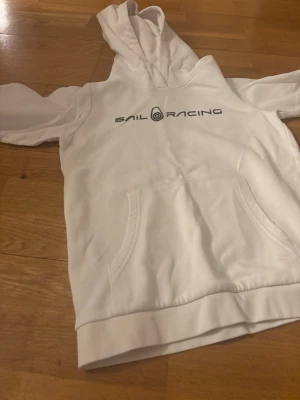 Vit hoodie från Sail Racing - Snygg vit hoodie från Sail Racing med svart logga tryckt över bröstet. Hoodien har en klassisk känguruficka framtill och en stor huva. Tillverkad i mjukt material som känns skönt mot huden. Perfekt för en avslappnad och sportig stil.