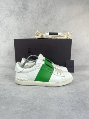 Valentino vita sneakers med grön detalj - Snygga vita sneakers från Valentino med en bred grön panel på sidan och klassiska nitar på sulan. Skorna har lågt skaft, vita snören och är tillverkade i läder. Perfekta för dig som vill ha en stilren men ändå unik look.