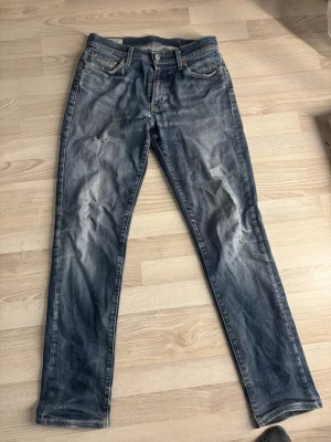 Levi's blå slitna jeans straight fit - Ett par klassiska blå jeans från Levi's med straight fit och snygga slitningar. Jeansen har fem fickor, knappgylf och tydliga whiskers-effekter framtill. Tillverkade i denim med en autentisk tvättad look och läderpatch bak i midjan. Mindre i midjan än vad storleken säger w32 L34