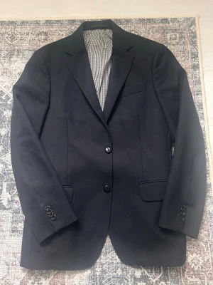 Mörkblå kavaj från Oscar Jacobson - Stilren mörkblå kavaj från Oscar Jacobson med klassisk passform och två knappar framtill. Kavajen har fickor med lock, bröstficka och snyggt rutigt innerfoder. Perfekt för en dressad look och tillverkad i ett mjukt tyg som känns lyxigt.