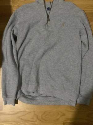 Grå halvzip tröja från Polo Ralph Lauren - Klassisk grå långärmad tröja från Polo Ralph Lauren med halv dragkedja och liten broderad logga på bröstet. Tillverkad i mjukt sweatshirtmaterial och har ribbade muddar vid ärmslut och nederkant. Perfekt för en avslappnad och stilren look.