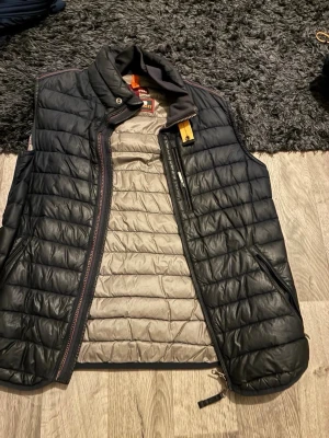 Svart dunväst från Parajumpers - Snygg svart dunväst från Parajumpers med quiltad design och gul detalj vid kragen. Västen har dragkedja, hög krage och praktiska fickor med dragkedja. Insidan är ljusgrå och materialet är lätt och värmande. Perfekt för lager-på-lager och streetwear.