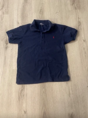 Mörkblå pikétröja från Ralph Lauren - Klassisk mörkblå pikétröja från Ralph Lauren med korta ärmar och krage. Tröjan har två knappar framtill och den ikoniska röda logotypen broderad på bröstet. Tillverkad i mjuk bomull för en bekväm känsla. Storlek M men i barnstorlek
