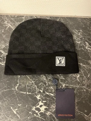 Svart mössa  - Snygg svart mössa. med diskret rutmönster och vikt kant. Märkt med logga framtill på en vit patch. Tillverkad i mjukt stickat material som känns lyxigt och stilrent. Perfekt accessoar för dig som gillar exklusiva detaljer.