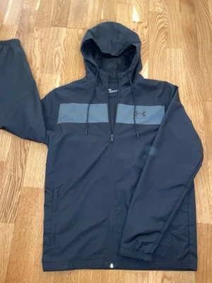 Svart vindjacka från Under Armour - Snygg svart vindjacka från Under Armour med huva och dragkedja framtill. Jackan har en grå panel över bröstet och Under Armour-logga på vänster sida. Tillverkad i lätt, vindtätt material med meshfoder och två sidofickor. Perfekt för blåsiga dagar. Helt ny och oanvänd.