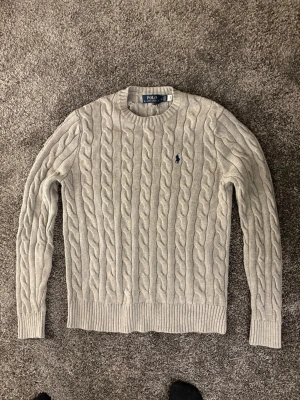 Ljusgrå kabelstickad tröja Polo Ralph Lauren - Kabelstickad ljusgrå tröja från Polo Ralph Lauren med rund halsringning och ribbade muddar. Tröjan har det klassiska broderade logotypen i mörkblått på bröstet och är tillverkad i mjukt material. Perfekt för en stilren och avslappnad look.