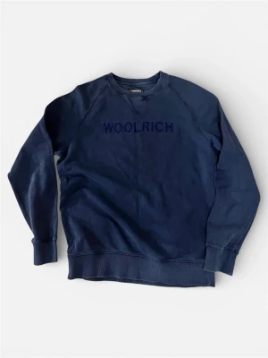 Blå sweatshirt från Woolrich - Mörkblå sweatshirt från Woolrich med rund halsringning och broderad logga på bröstet. Tröjan har långa ärmar, ribbade muddar och avslappnad passform. Perfekt för en casual och stilren look.