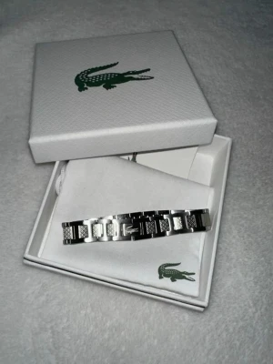 Silverfärgat armband från Lacoste - Snyggt armband i silverfärgad metall från Lacoste med klassisk krokodil-logga på länkarna. Armbandet har en stilren och modern design med detaljerade mönster på vissa länkar. Perfekt accessoar för dig som gillar en clean och sportig look.