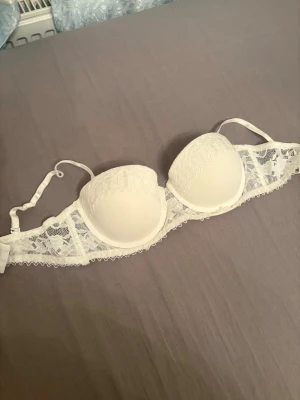 Vit spets-bh med vadderade kupor - Snygg vit bh med vadderade kupor och fina spetsdetaljer. Axelbanden är justerbara och ryggen har hakknäppning. Perfekt för dig som gillar en feminin och stilren look. Materialet är en mix av spets och slät textil för extra komfort.