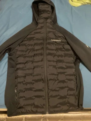 Svart jacka från Peak Performance - Snygg svart jacka från Peak Performance med huva och dragkedja framtill. Jackan har quiltad design på fram- och baksidan, samt två dragkedjeförsedda fickor. Perfekt inför våren och har en sportig look.